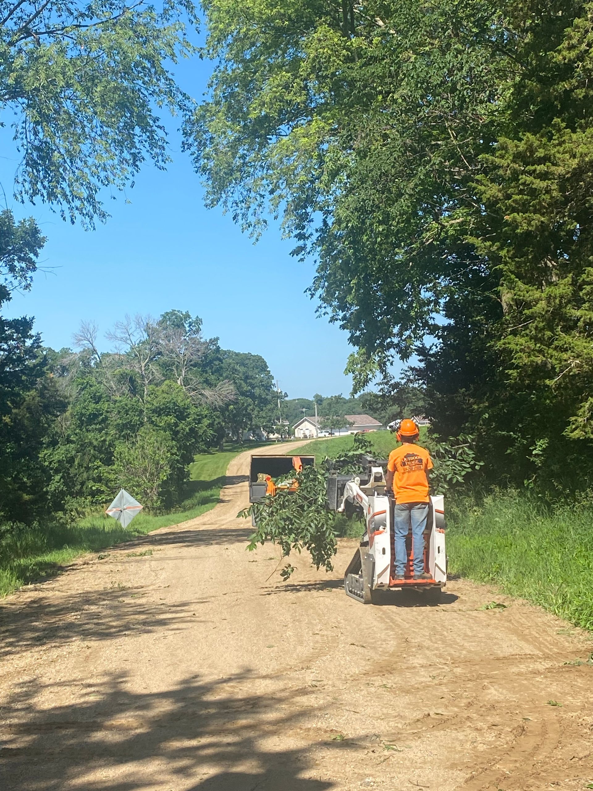 People walking on a sunny dirt road under trees, with one wearing an orange shirt in front.