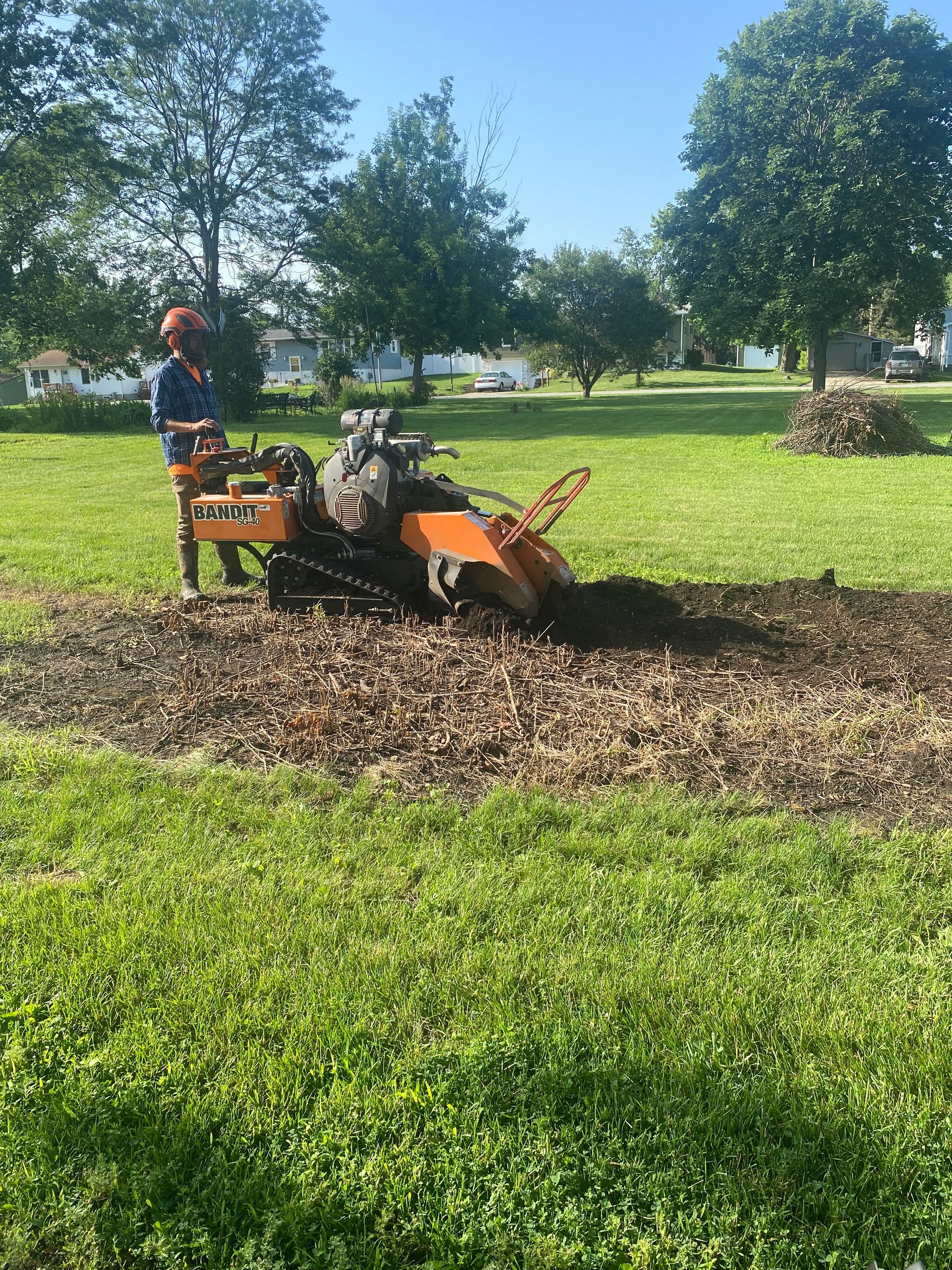Person operating an orange machine in a grassy park, clearing a patch of mulch under trees.