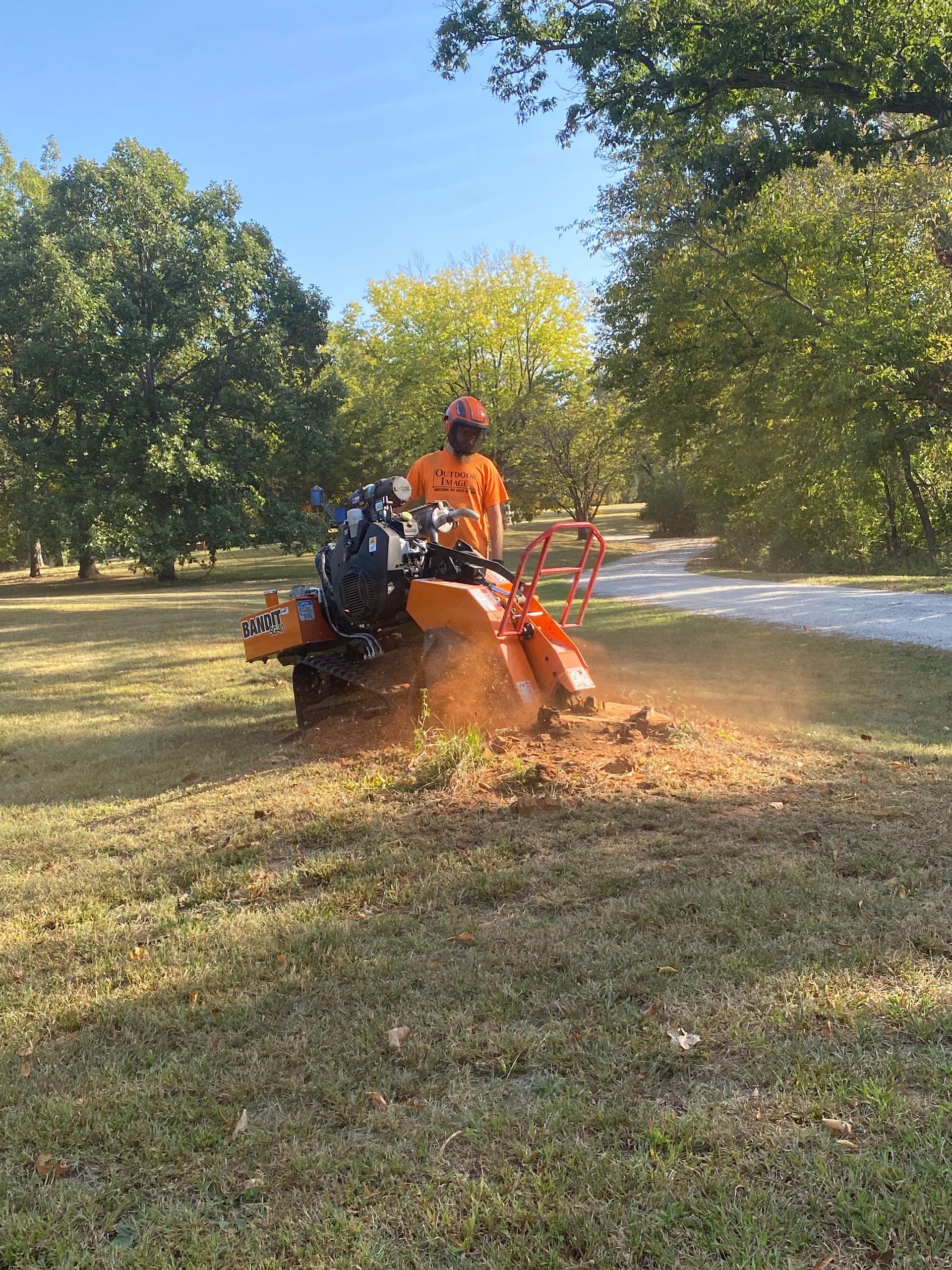 Person in orange gear using a mower to clear leaves on a grassy field with trees nearby