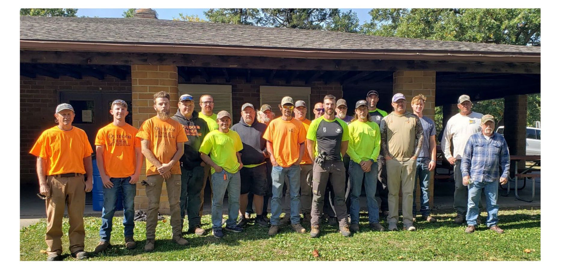 Group of people standing outdoors in front of a wooden pavilion, some wearing bright orange and yellow shirts.