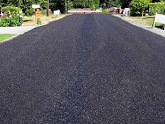 Asphalt sealcoating — Lane County, OR — Miles Asphalt Solutions A freshly paved, black asphalt driveway stretches forward between green lawns and landscaping on a sunny day.