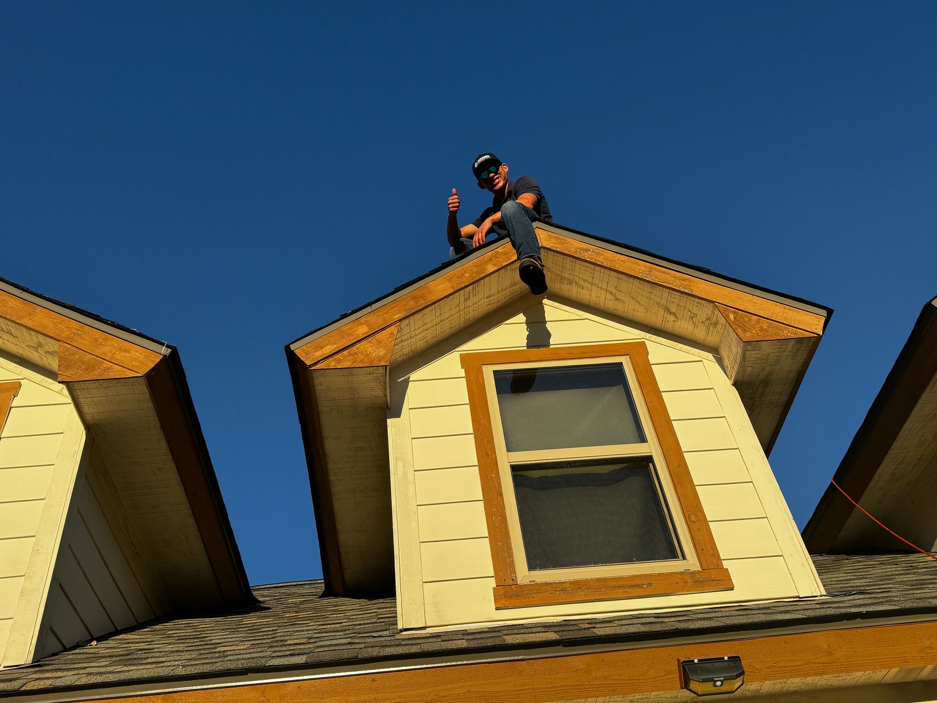 Person sits on a roof peak, giving a thumbs up. Blue sky backdrop. Light yellow siding, brown trim, and grey roof.