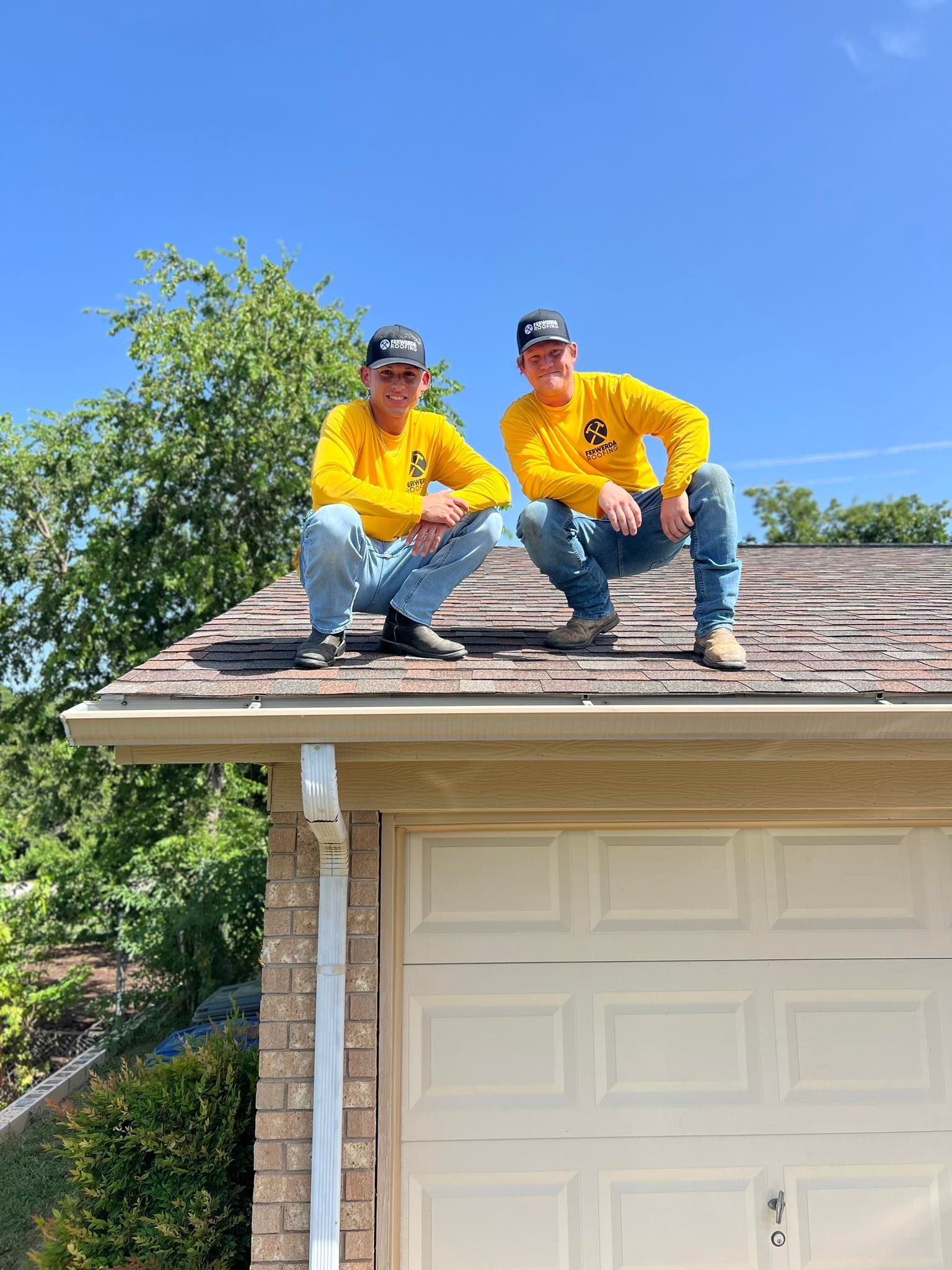 Two people in yellow shirts and caps crouched on a roof, above a garage door, under a blue sky.
