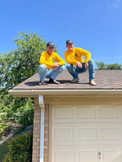 Two people in yellow shirts and caps crouched on a roof, above a garage door, under a blue sky.