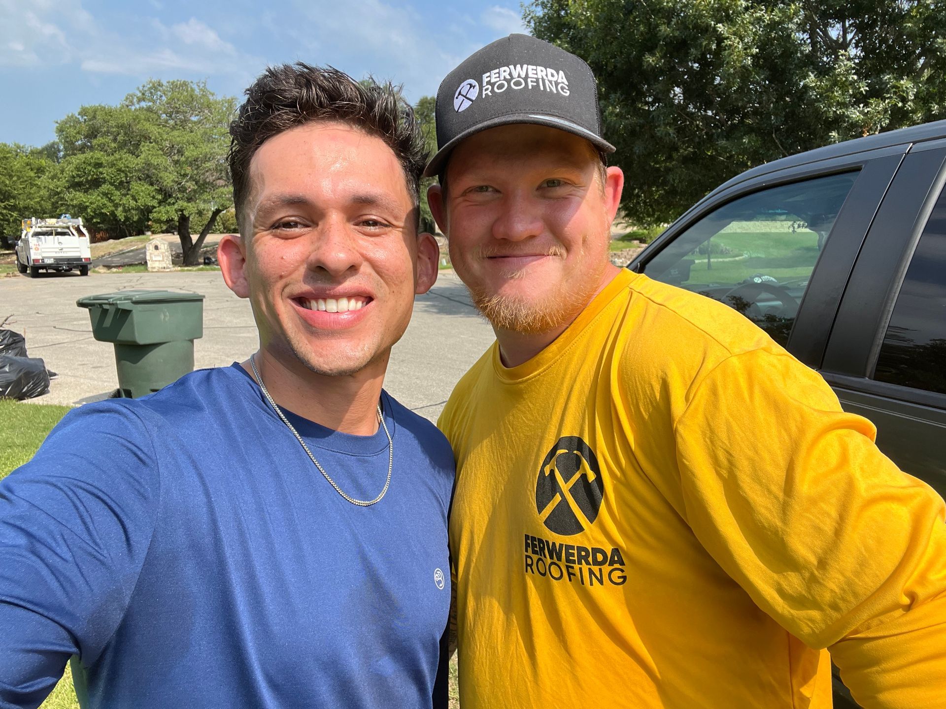 Two men smiling, one in a blue shirt, the other in a yellow shirt and cap, standing outside next to a truck.