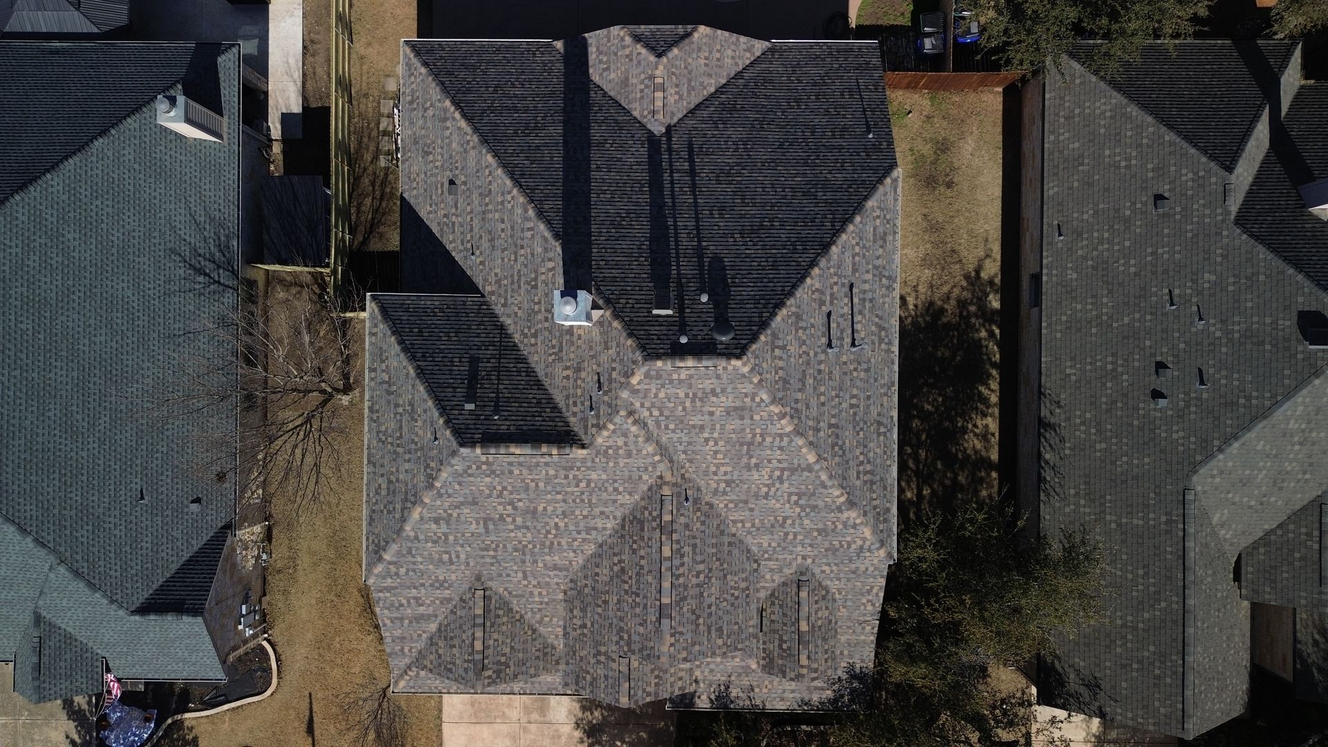 Overhead view of a house with a complex, dark gray roof. Other houses and trees surround it.
