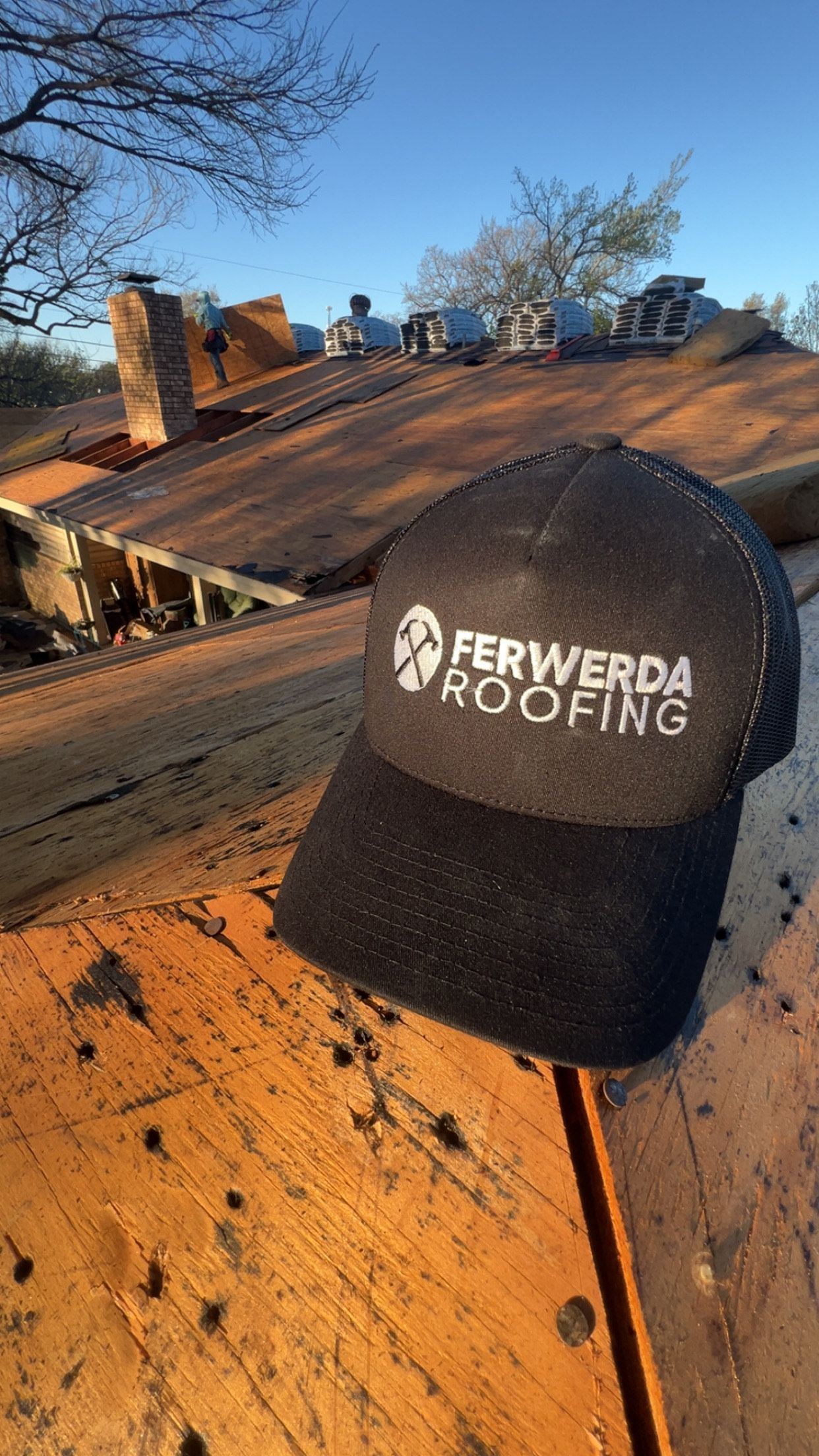 Black Ferwerda Roofing hat on a weathered wooden roof with chimney and blue sky background.