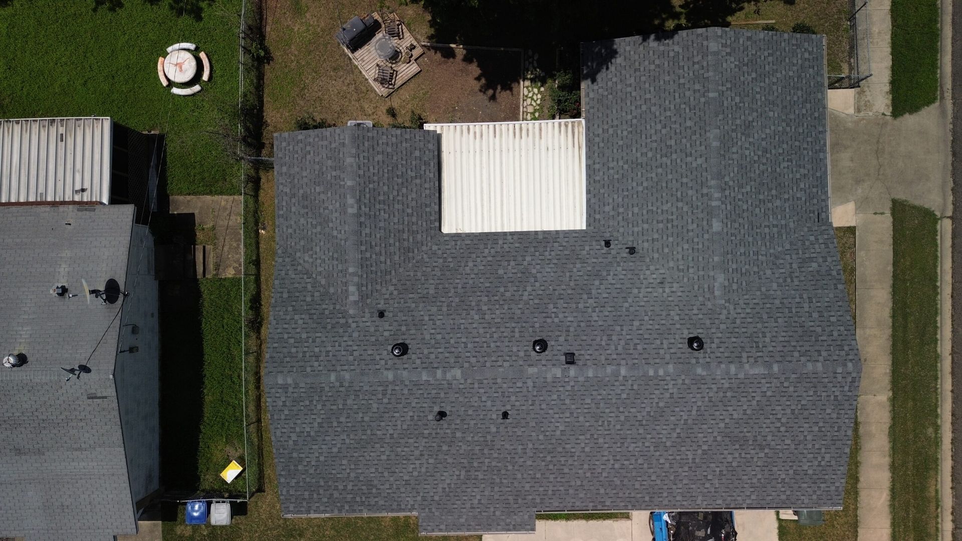 Overhead view of a dark gray shingled roof with a white section, next to green grass and a house.