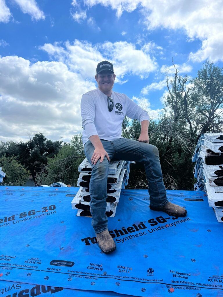 Man sits on shingles on a rooftop, wearing work attire. Blue sky and clouds are in the background.