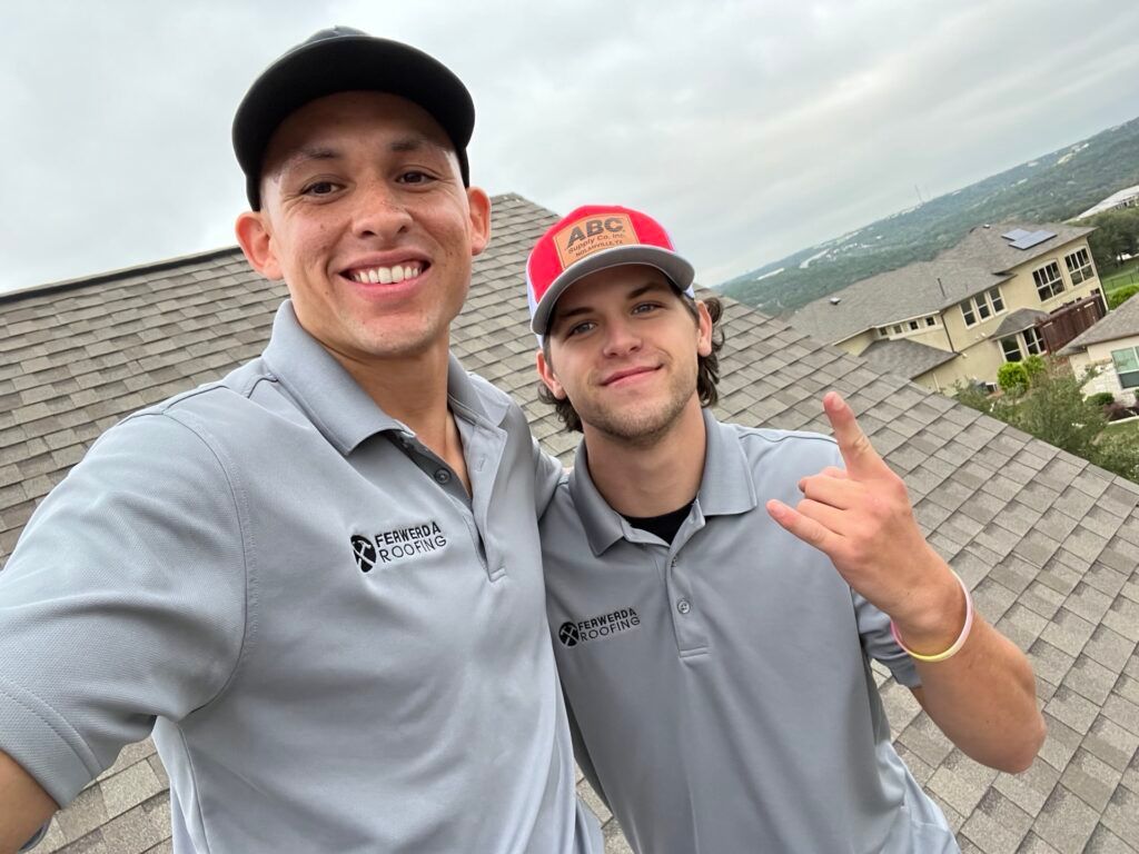 Two men smiling on a rooftop wearing gray polo shirts and baseball caps, one flashing a hand gesture.