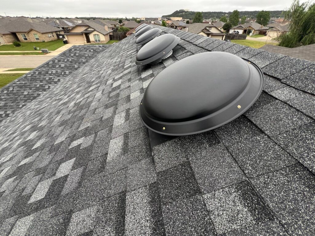 Gray asphalt shingle roof with three black dome-shaped skylights. Residential neighborhood in the background.