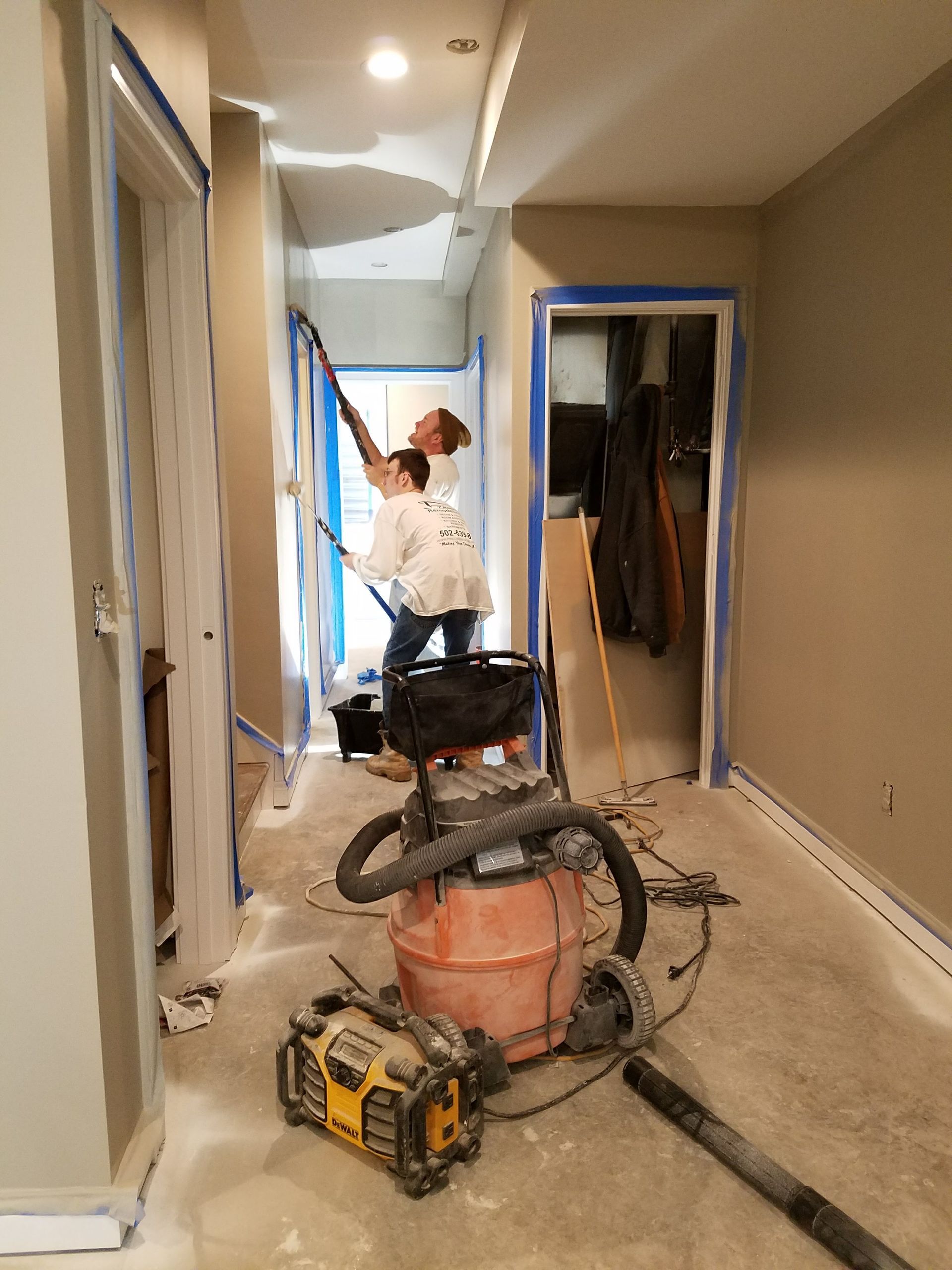 A man is painting a wall in a hallway next to a vacuum cleaner.