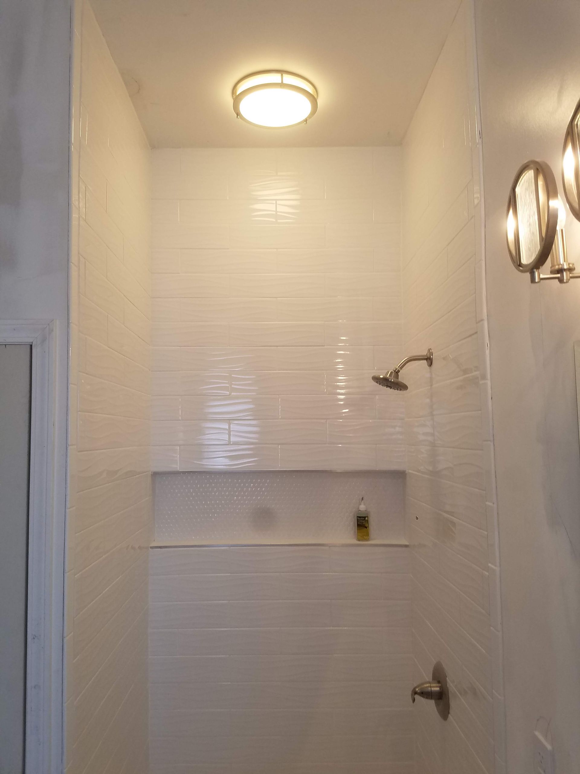 A bathroom with white tiles and a shower with a light on the ceiling.