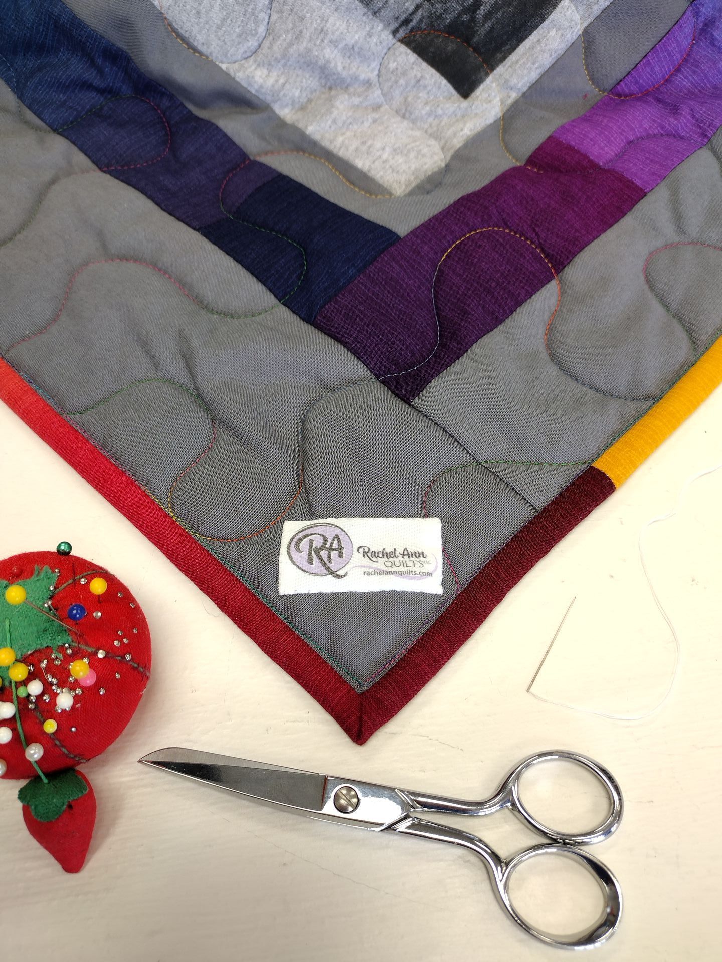 The corner of a quilt with the Rachel Ann Quilts label, a small scissors, and a Tomato pincushion with pins and needle.