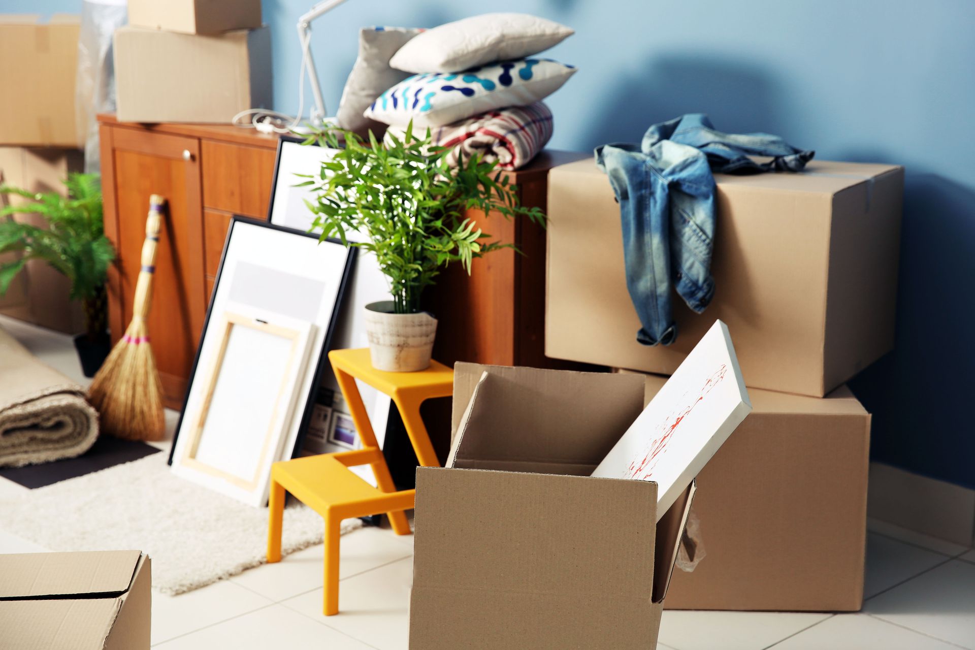 A room filled with cardboard boxes, packed items, a broom, a potted plant, and furniture, suggesting a moving process.