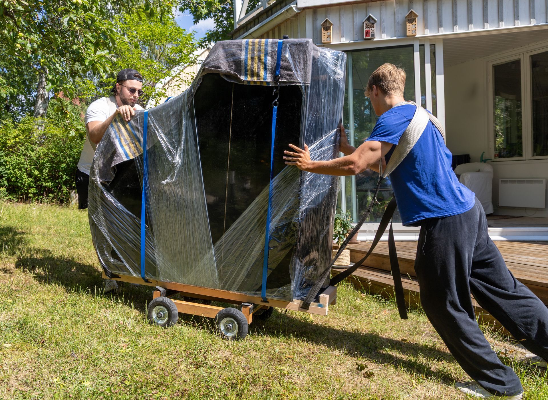 Two people move an upright piano wrapped in plastic and blankets on a wooden dolly across a grassy lawn towards a house.