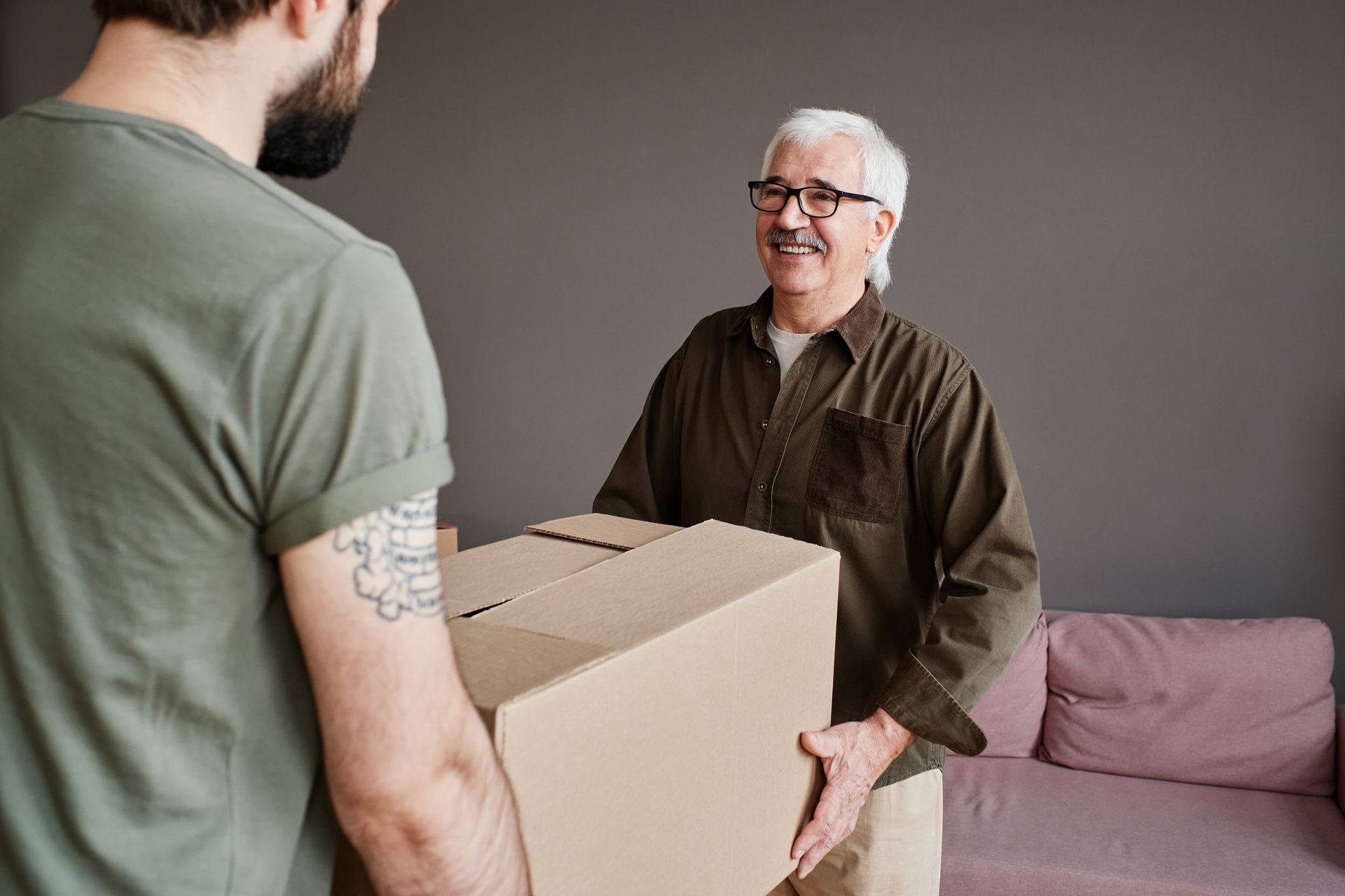 Two people in a living room, smiling as they jointly lift and carry a large brown cardboard moving box.