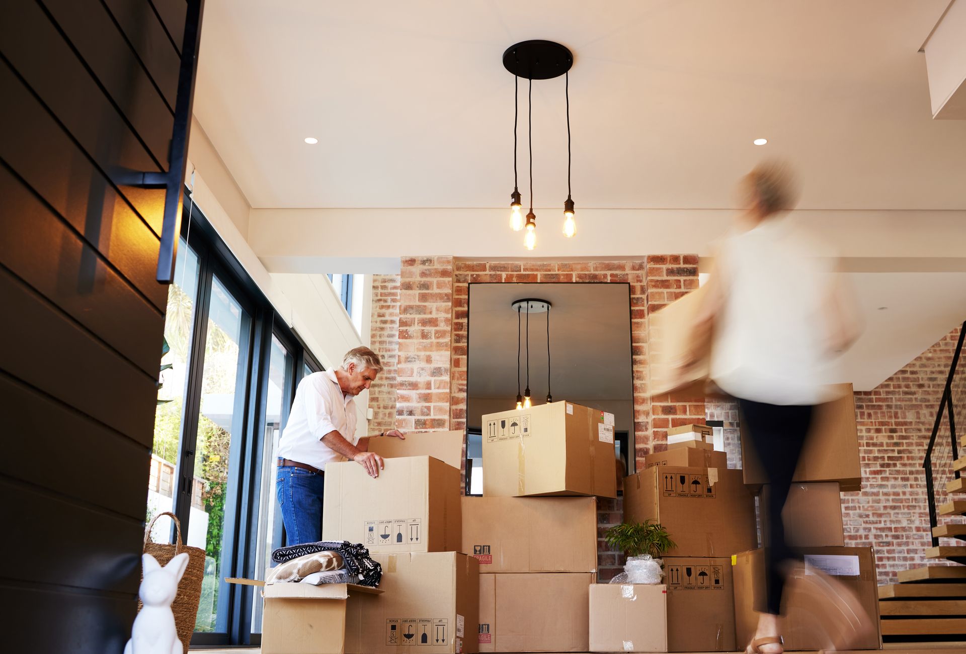 A person packs boxes while another walks past in a home with exposed brick walls, preparing for a move.