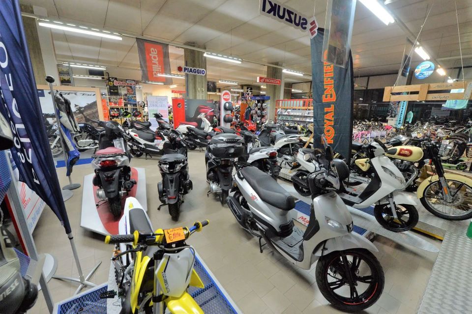 Salone motociclistico con vari scooter e moto in esposizione. Bandiere Suzuki, Honda e Royal Enfield pendono dal soffitto.