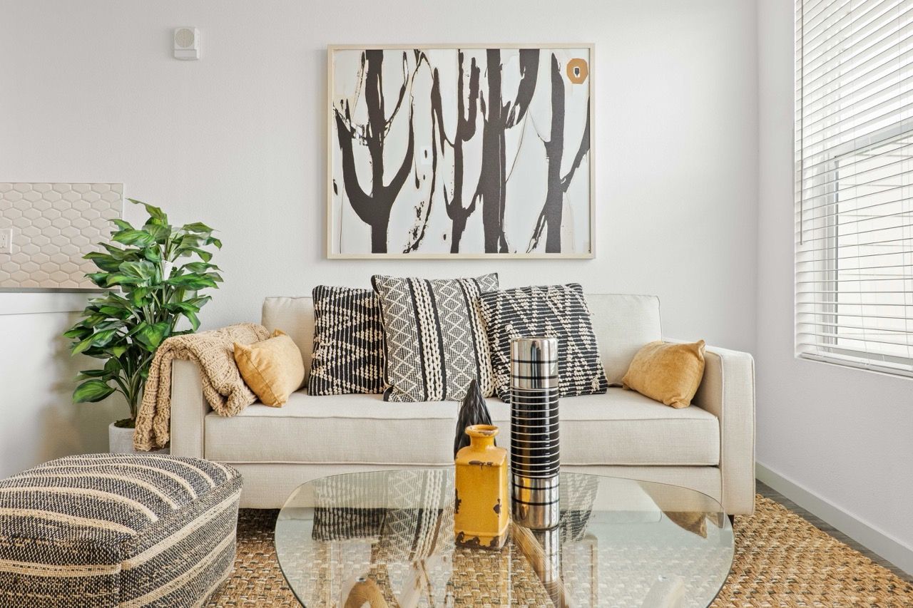 Living room with beige sofa, patterned cushions, glass coffee table, plant, and abstract wall art.