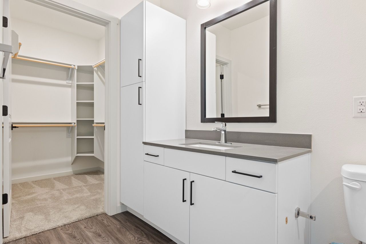 Modern bathroom vanity with white cabinetry, gray countertop, and a walk-in closet visible through an open doorway.