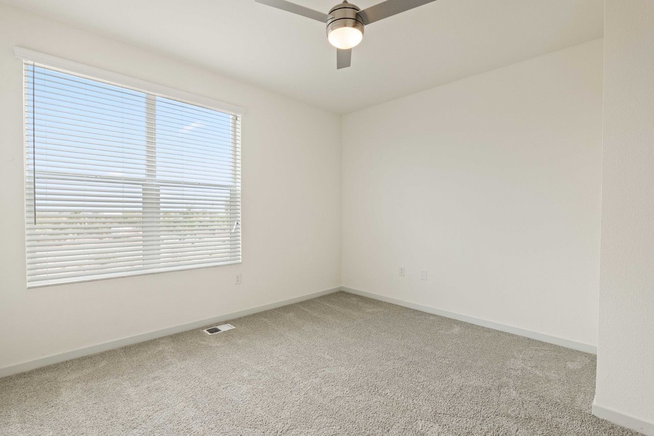 Empty carpeted bedroom with a large window and white blinds; ceiling fan overhead.