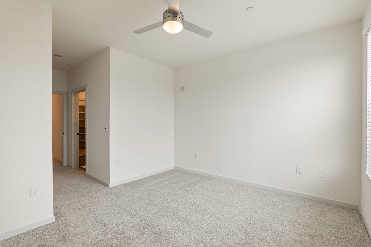 Empty bedroom with white walls, beige carpet, and a ceiling fan.