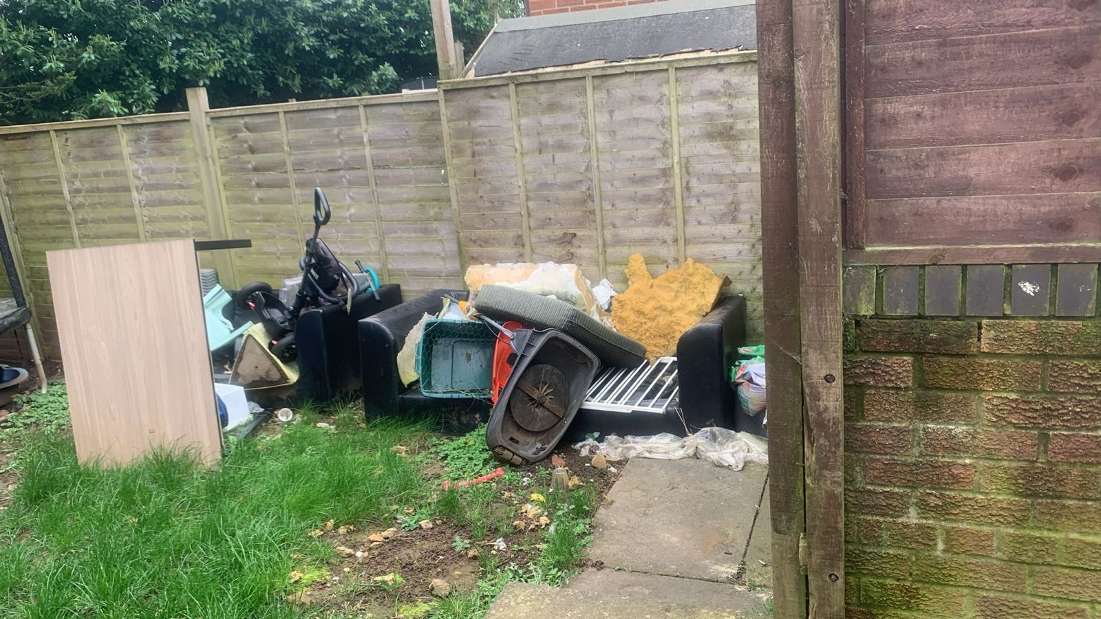 A backyard garden scene with a collection of discarded furniture, including a sofa, foam padding, and wooden boards.