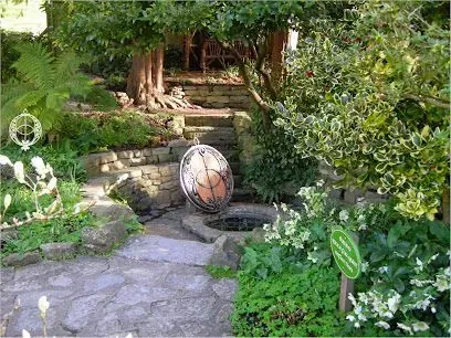 Stone garden path leading to a well, with steps and surrounding lush greenery, including trees and flowering plants.