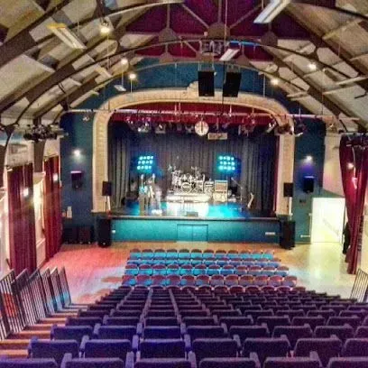 Auditorium with stage and blue seating. Stage has a band setup. Teal walls, red curtains, wood floors.