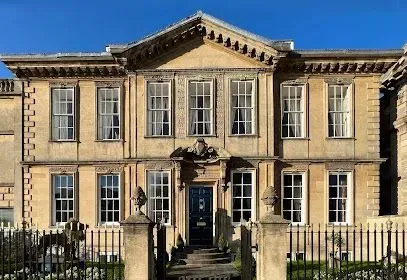 Two-story stone building with symmetrical facade, white-framed windows, dark door, and decorative trim under a blue sky.