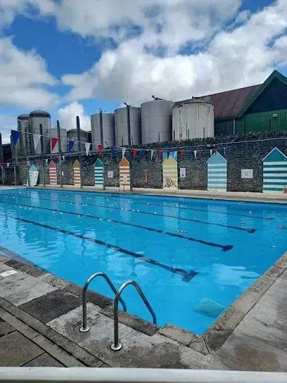 Outdoor swimming pool with blue water and lane markers, surrounded by colorful changing huts under a cloudy sky.