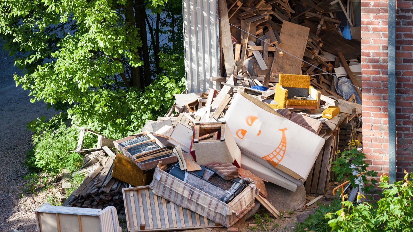 Pile of discarded furniture and debris outside, next to a brick building and green foliage.