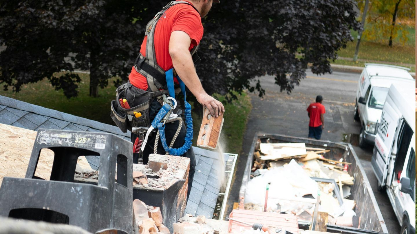 Roofer in a safety harness on a roof, tossing brick into a dumpster. Another worker nearby.