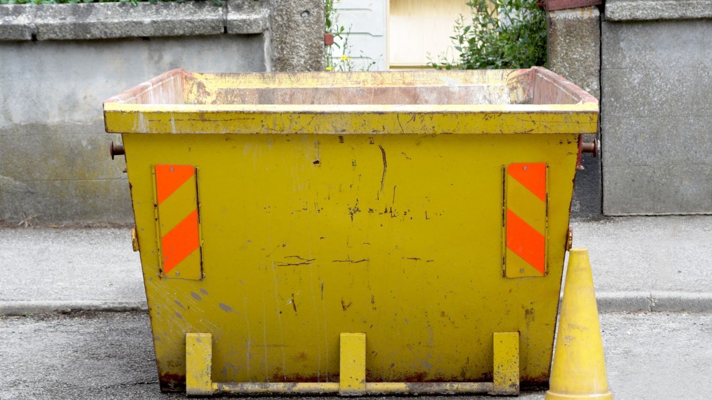 Yellow dumpster with orange reflective panels and a traffic cone on a sidewalk.