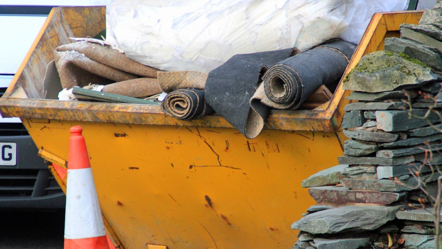 Yellow skip filled with rolled carpet and a traffic cone. Stone wall in the foreground.