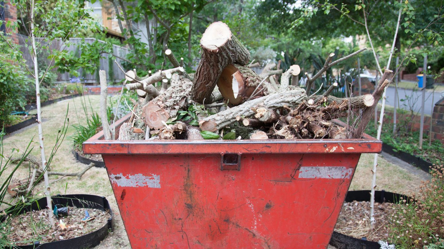 Red dumpster overflowing with freshly cut branches and logs in a garden setting.