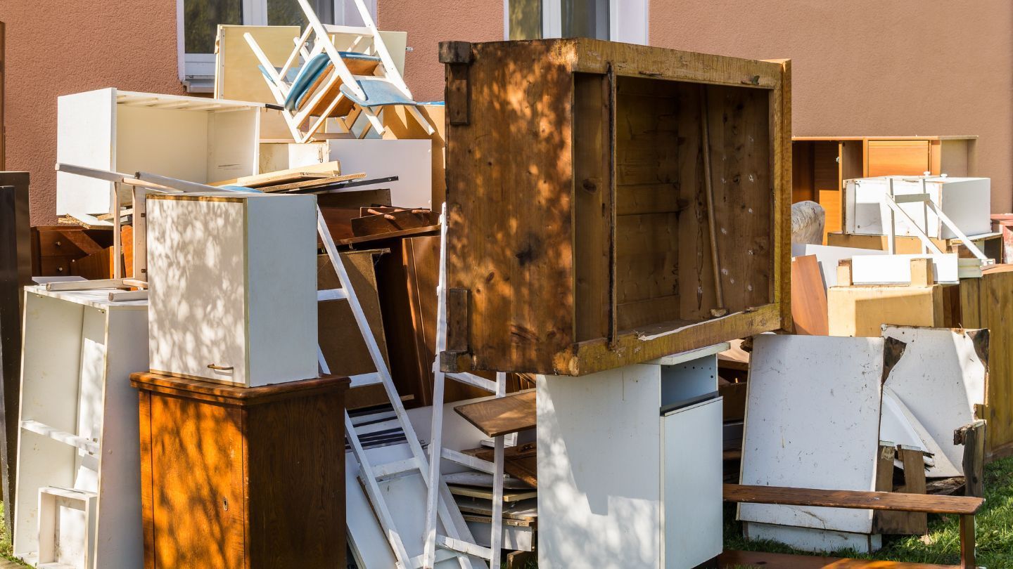 Pile of discarded wooden furniture outdoors, including cabinets, drawers, and a ladder, against a building.
