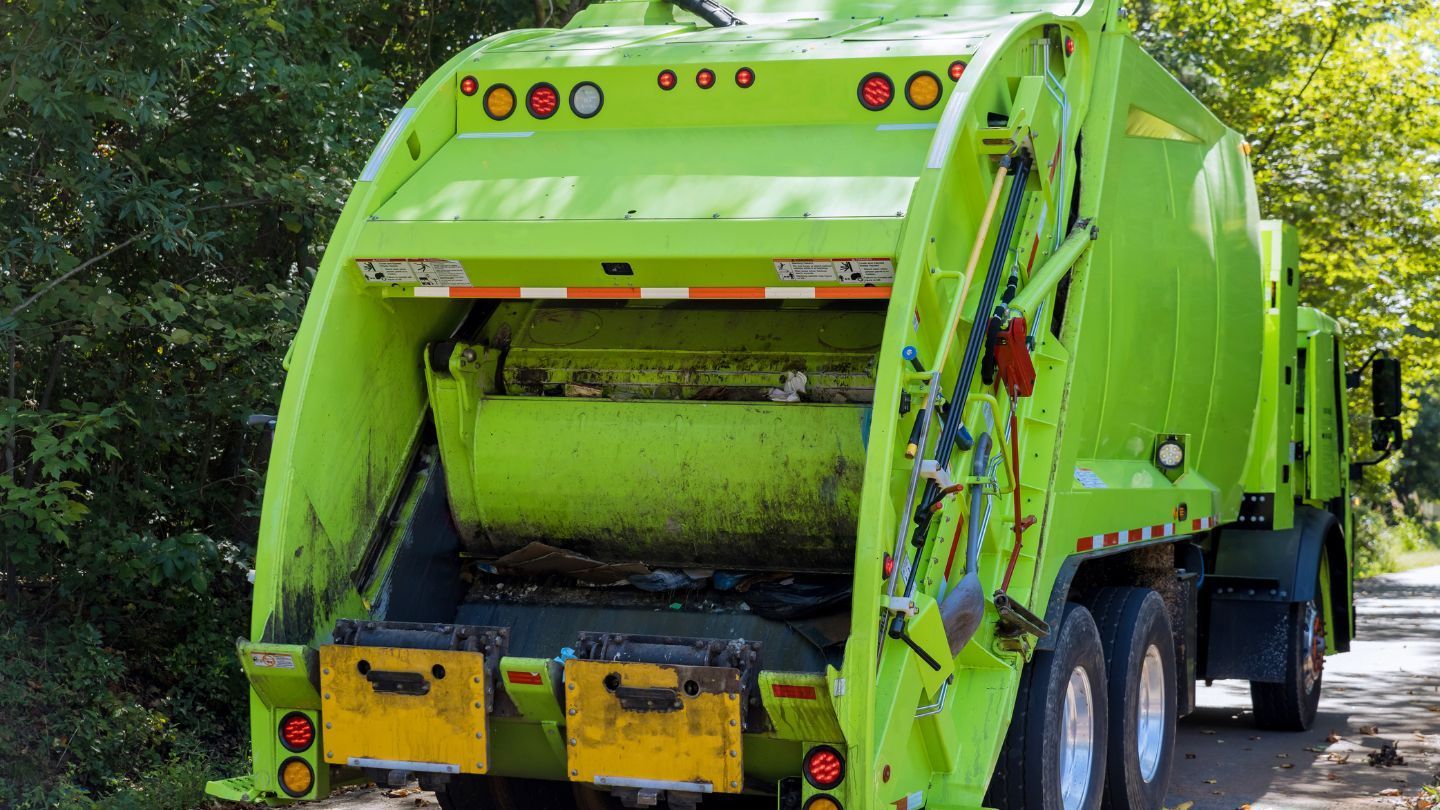 Green garbage truck on a road, with the back open, filled with trash.