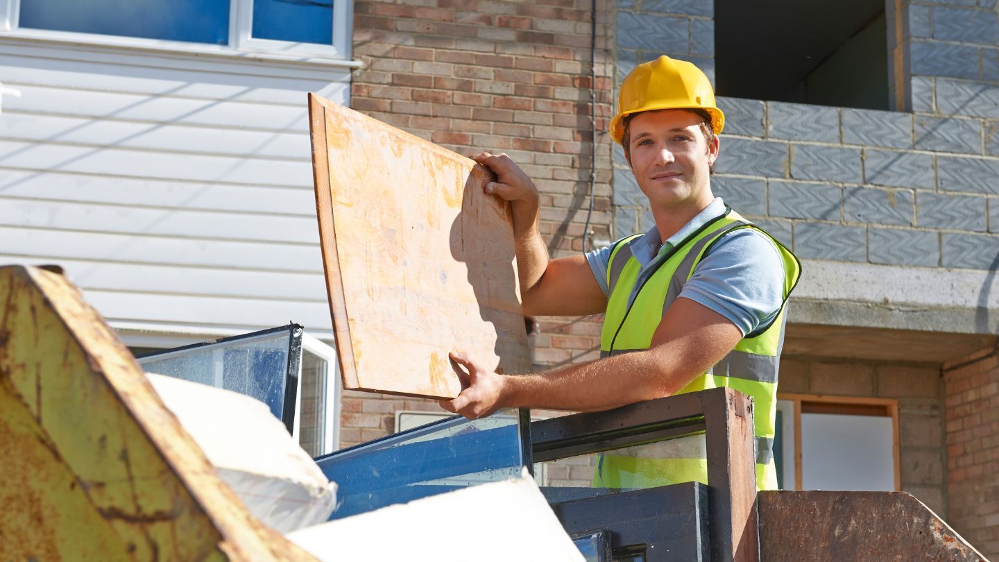 Construction worker in hard hat and vest placing wood into a dumpster.