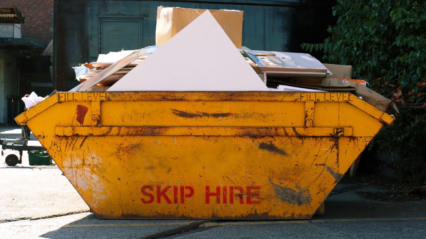 Yellow skip filled with construction debris, including cardboard and a white panel, outdoors.
