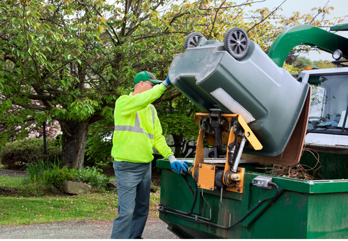 Garbage truck operator emptying a green trash can into a compactor. The worker wears a yellow safety vest.