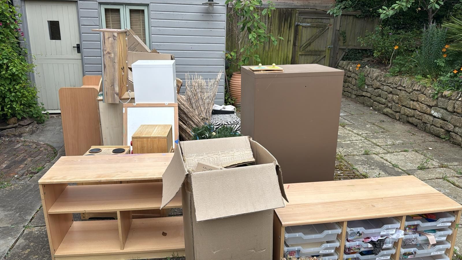 Stacks of boxes and wooden furniture piled outdoors in a backyard beside a house.