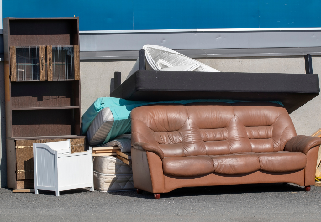 Pile of discarded furniture: brown leather sofa, bookcase, bed frame, and other items.