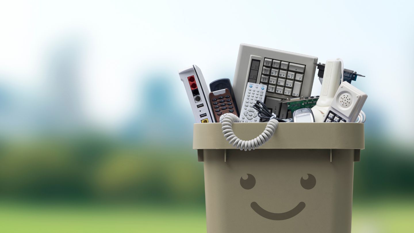 A bin overflowing with old electronics against a blurry outdoor background; the bin has a smiley face.