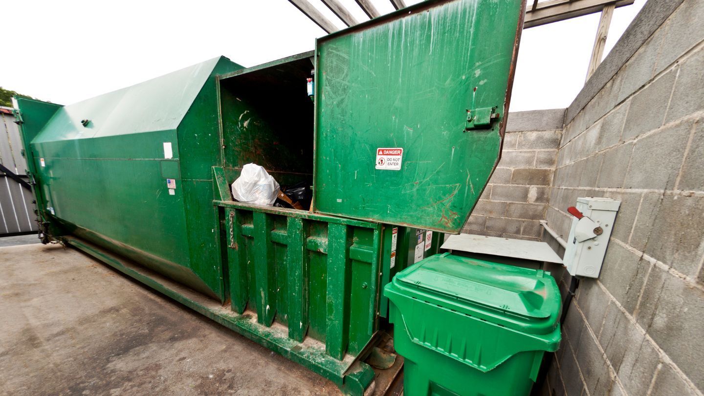 Green industrial trash compactor with open door, next to a green trash bin and brick wall.