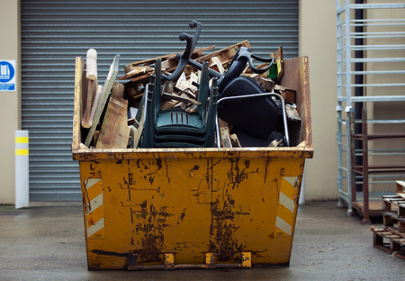 Yellow dumpster overflowing with debris, including furniture, against a gray building and pallets.