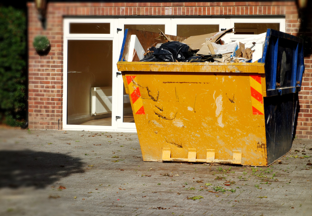 Yellow and black dumpster overflowing with debris in front of a brick building with open windows.