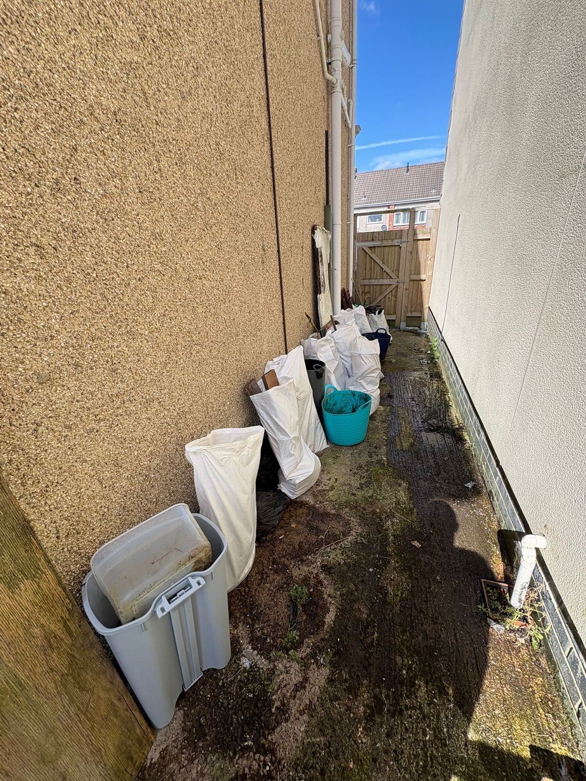 A narrow alleyway between two textured walls features several white storage bags, a grey bin, and a blue bucket.