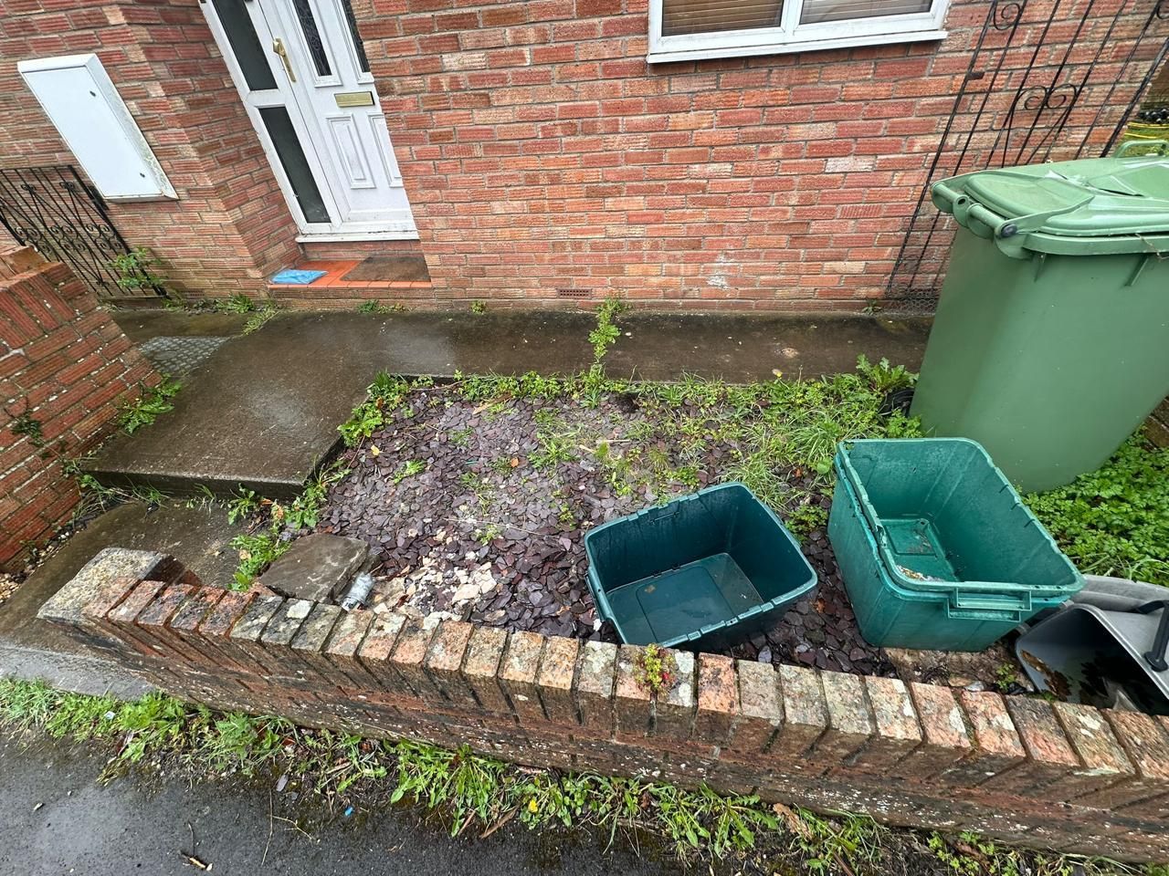 A small garden bed with two empty green plastic bins in front of a brick house with a white door and green wheelie bin.